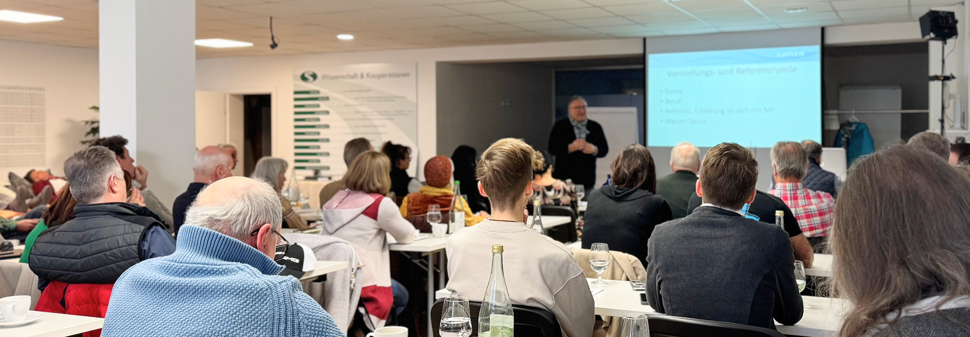 Publikum sitzt in einem Seminarraum und hört einem Vortragenden vor Leinwand zu